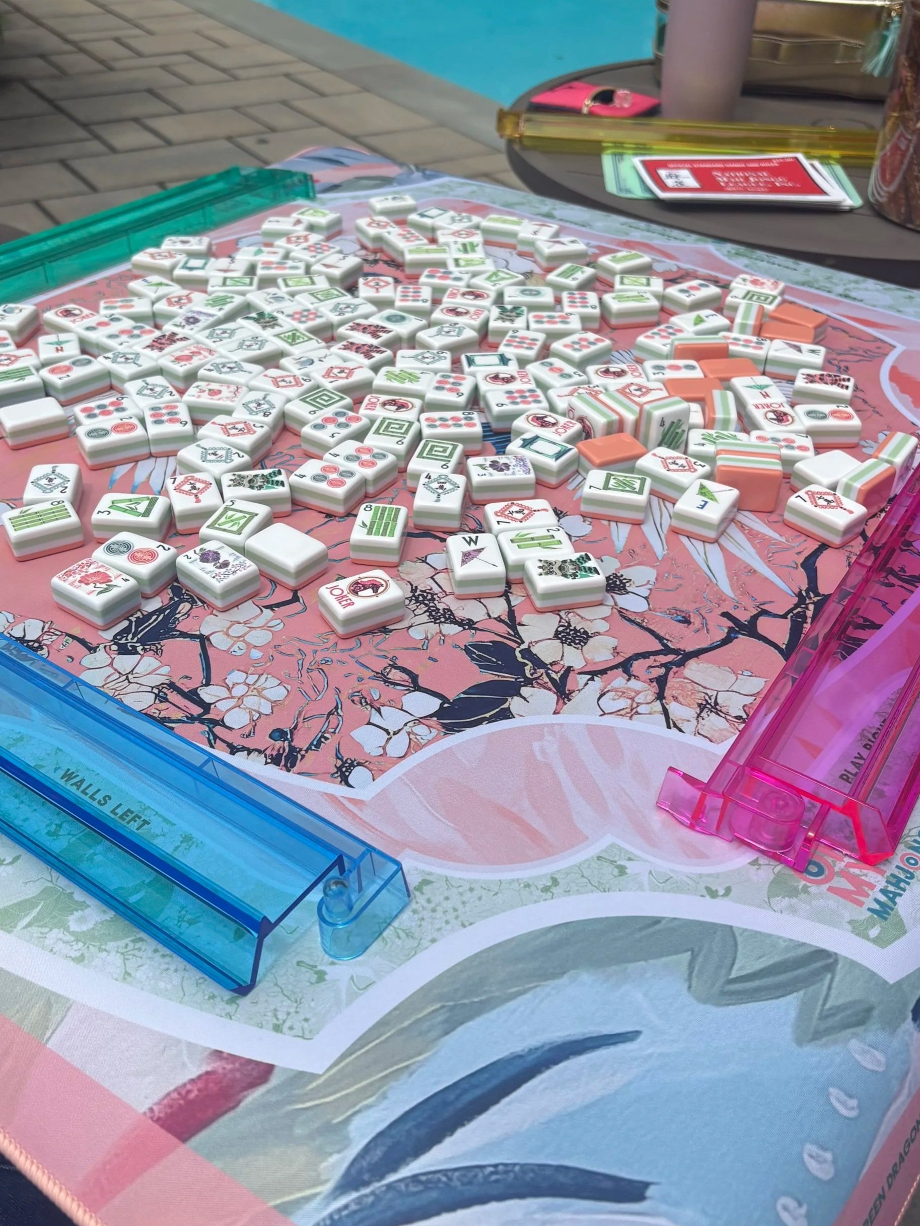 A colorful Mahjong game set up on a pink floral patterned table, with tiles scattered across the surface, two plastic racks (blue and pink) on opposite sides, and some items like a pink phone case, a pen, and folded papers on the side.