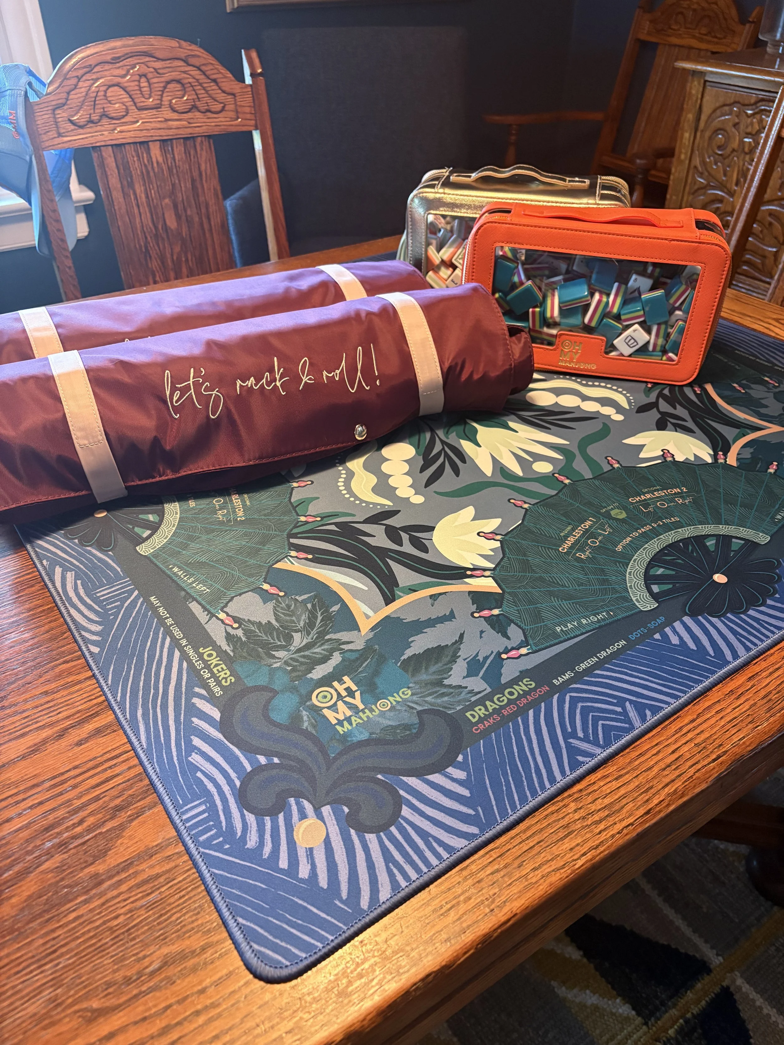 A table with a colorful Mahjong game mat featuring a dragon design, two transparent bags filled with Mahjong tiles, and a rolled-up bag with the phrase "let's pack & roll!" written on it.