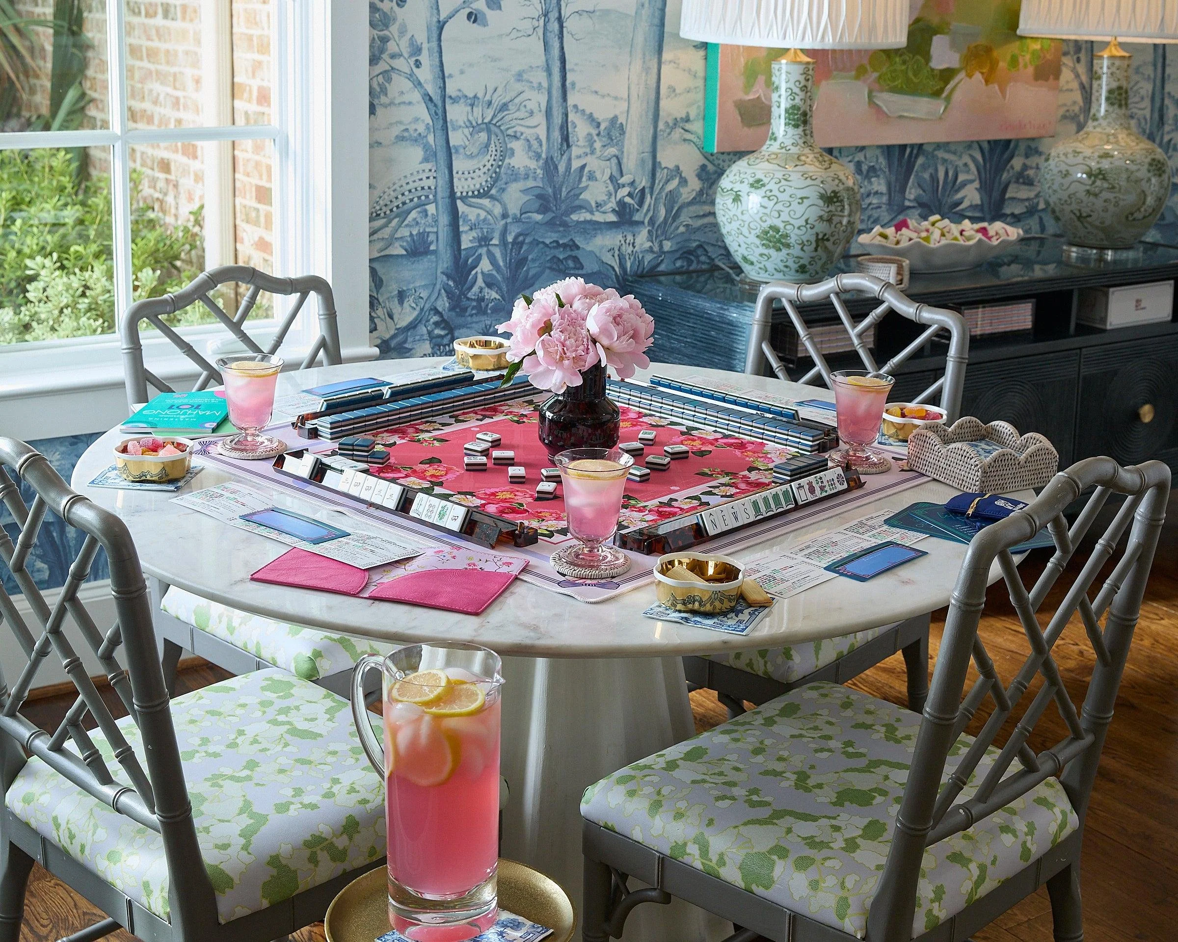 A dining room with a round table covered with a pink floral tablecloth, surrounded by chairs with green patterned cushions. The table has a central black vase with pink flowers, several cards or clips, and pink glasses with lemon slices. A tall pink drink with lemon slices is in the foreground. In the background, there are large decorative vases with green and white patterns, a sideboard, and a blue and white mural of a landscape with trees and mountains.