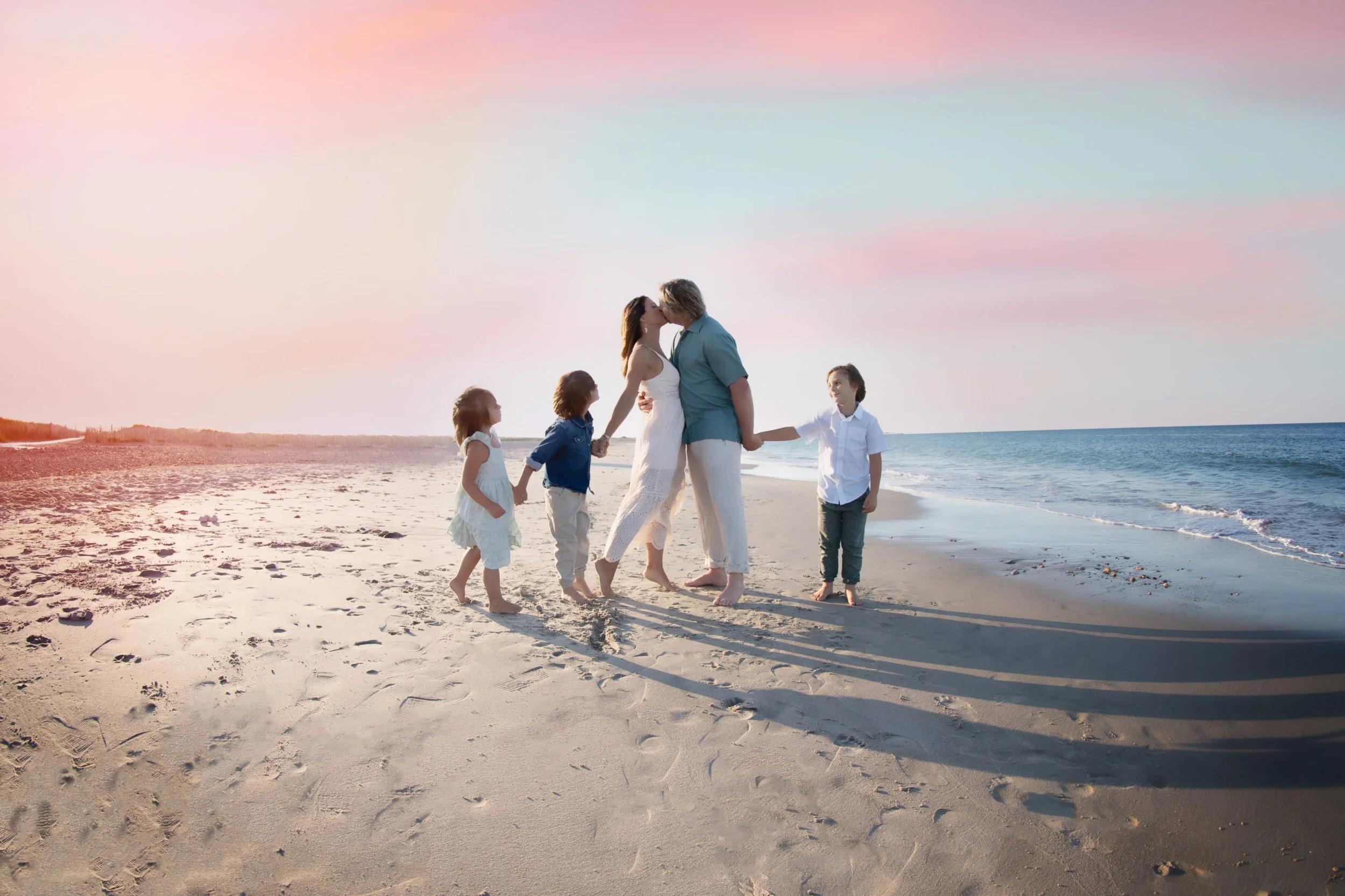 A family of five standing on the beach, holding hands and kissing during sunset. The sky is painted with pastel pink, blue, and purple colors, with the ocean in the background. The sand is dotted with footprints.