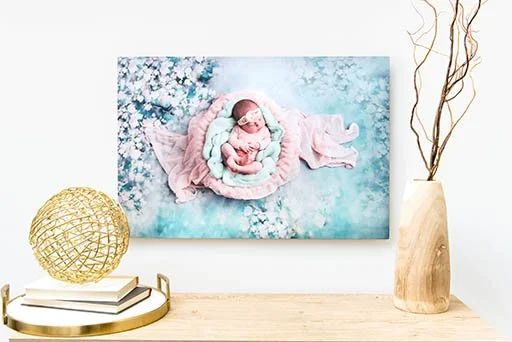 A framed photo of a newborn baby wearing a pink hat, lying on a pink blanket with a floral background, displayed on a white wall.