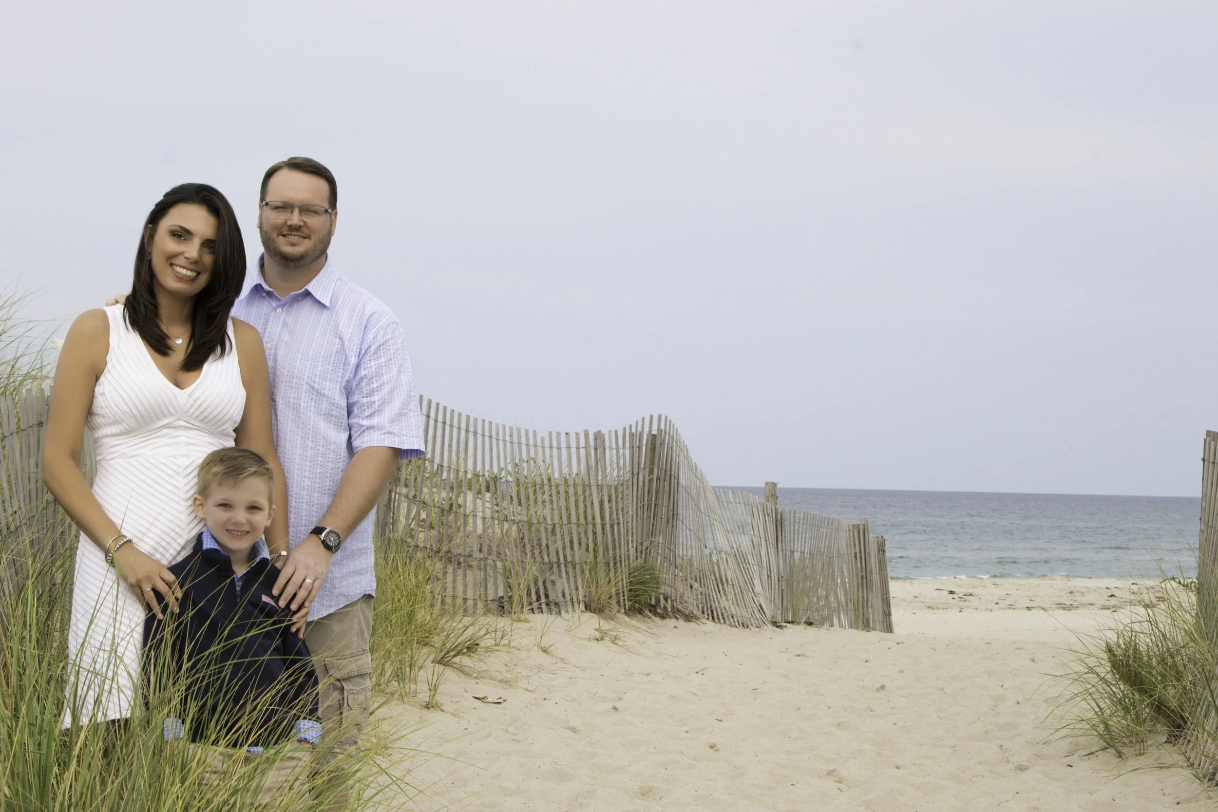 A family of four at the beach: woman, man, and two young boys, standing on sand with grassy dunes and a wooden fence behind them, ocean and cloudy sky in the background.