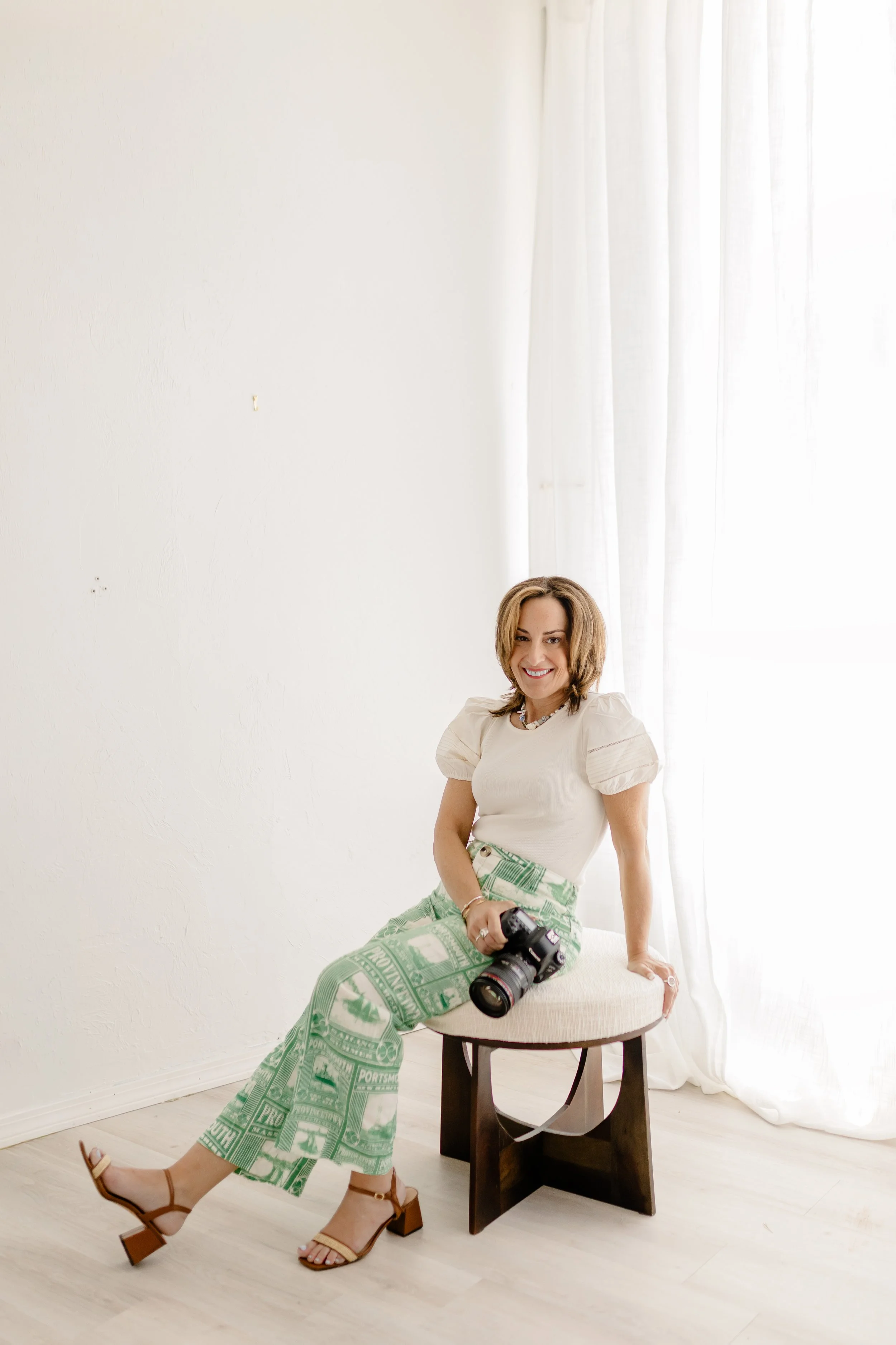 A woman with shoulder-length brown hair sitting on a small wooden stool in a bright room with white walls and curtains, smiling at the camera. She wears a cream-colored top, patterned green and white pants, and tan sandals. A camera rests on her lap.