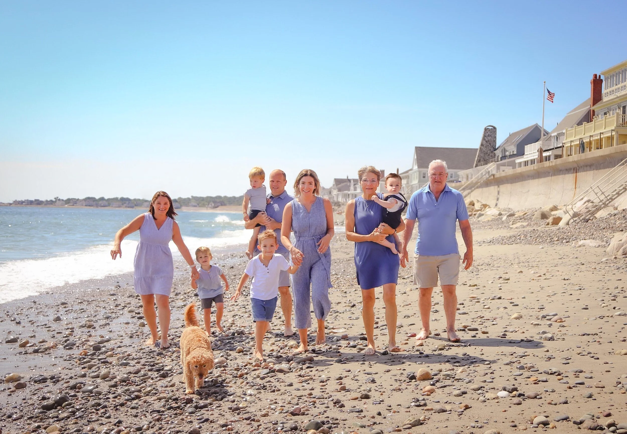 A group of people walking on a pebbled beach with houses and an American flag in the background. The group includes children, adults, and a dog.