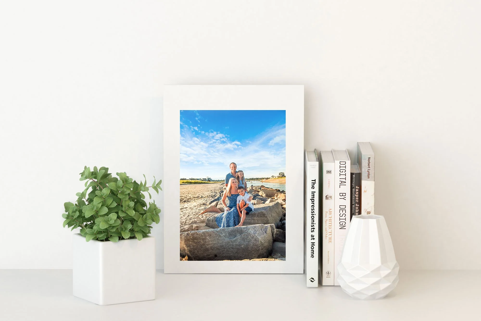 A white shelf with a framed photograph of a family at the beach, a green potted plant, and a row of books and a white geometric vase.