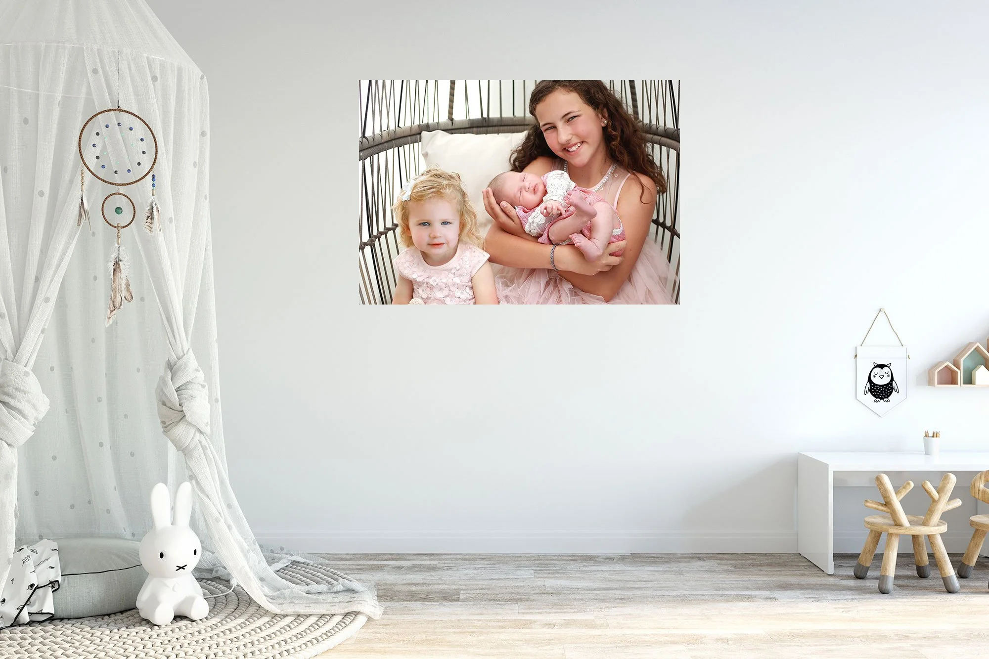 Family portrait on wall showing a smiling woman holding a newborn baby and sitting next to a young girl, all in a bright, modern nursery.