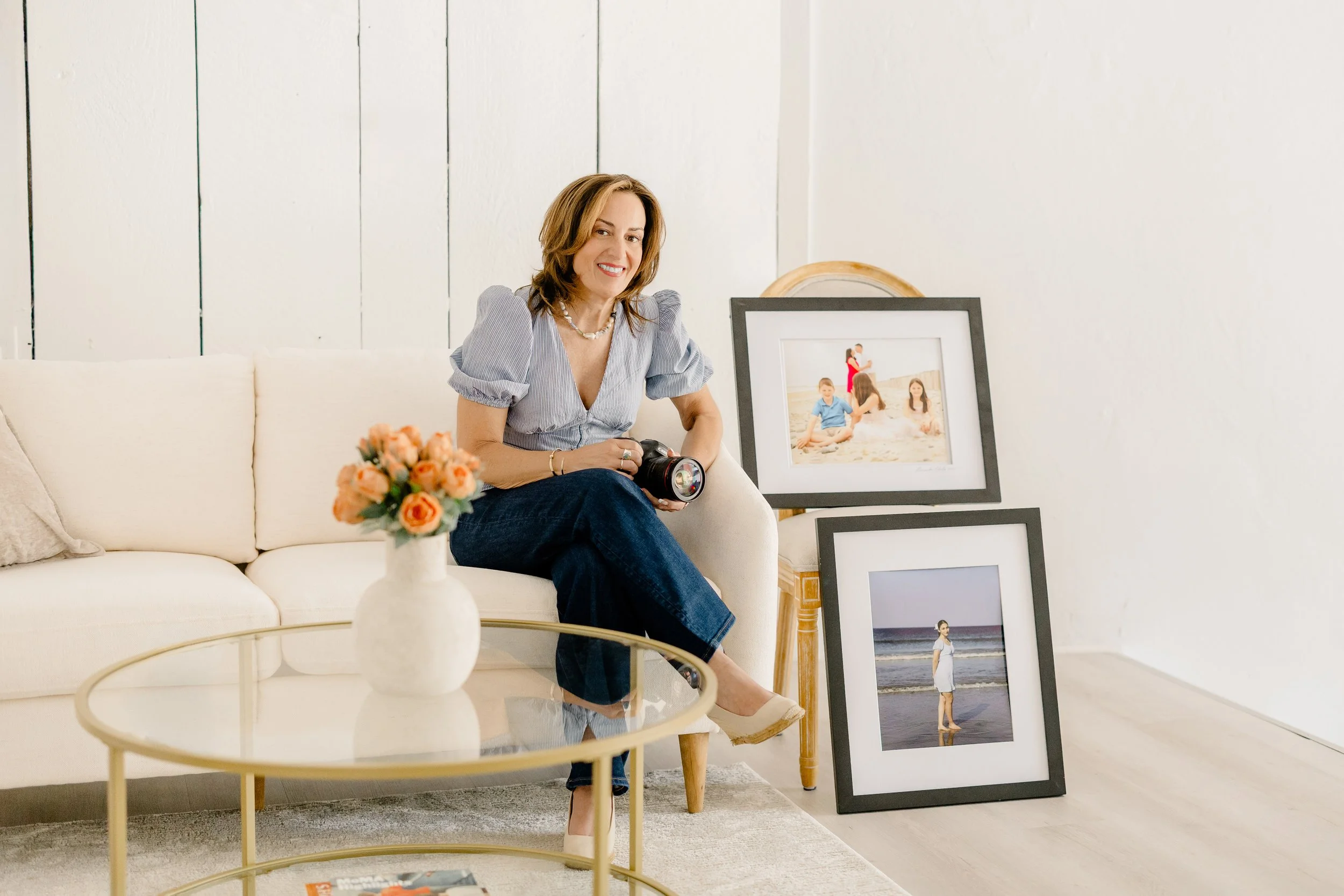 A woman sitting on a white couch in a living room, holding a camera, surrounded by framed photographs on the floor and a small round glass table with a vase of orange roses.
