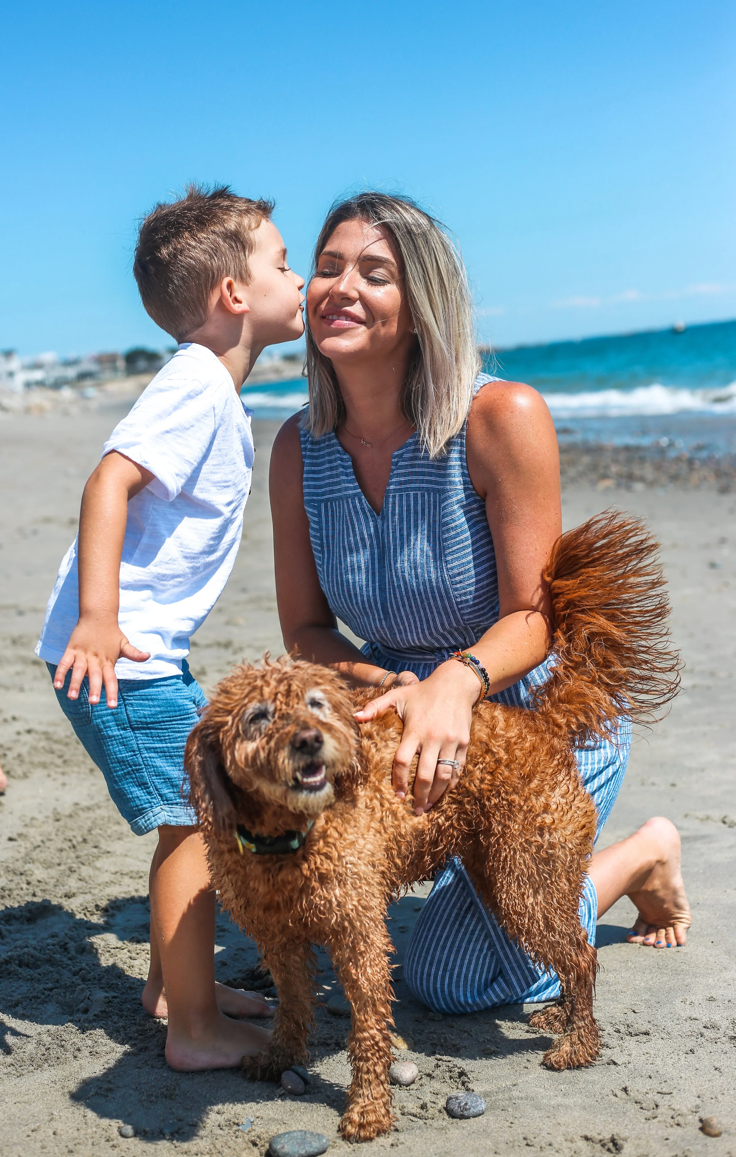 A woman and a young boy on the beach with a brown curly-haired dog, both smiling. The boy is kissing the woman on the cheek, and the woman is cuddling the dog. The beach has waves and a clear blue sky.