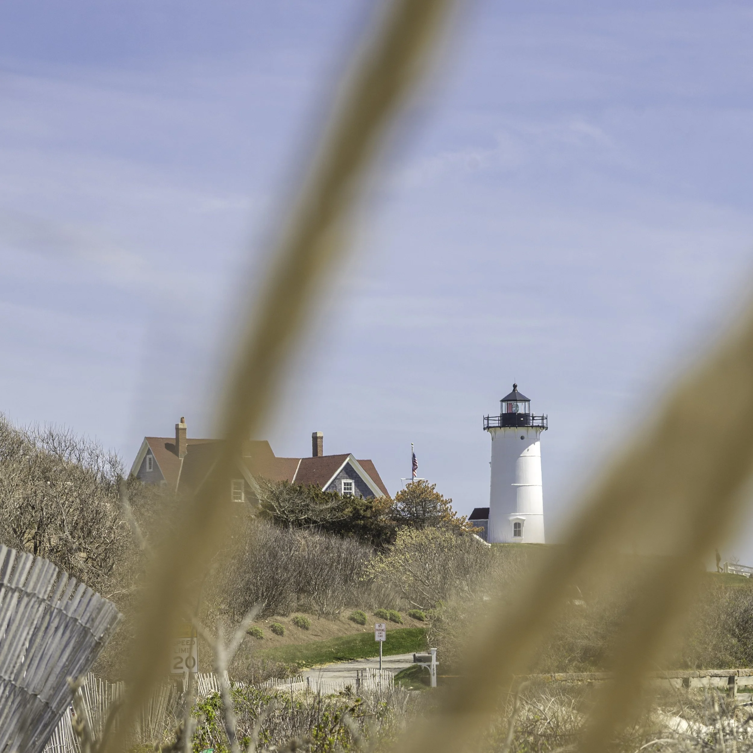 A lighthouse seen through a chain-link fence with a residential neighborhood in the background under a clear blue sky.