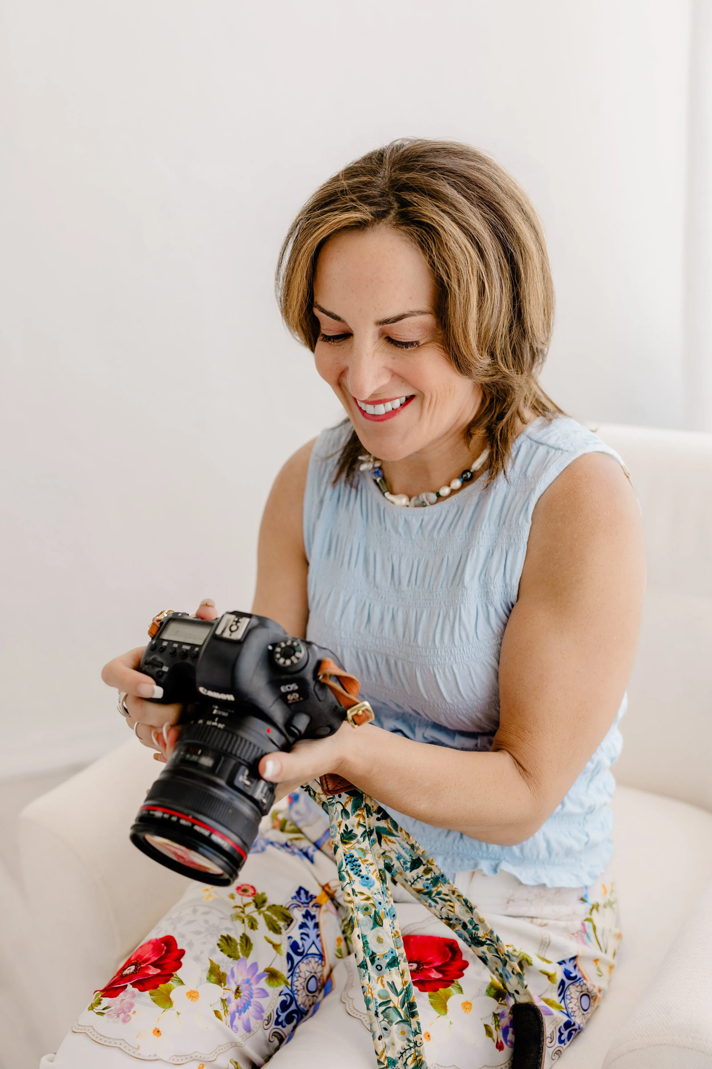 A woman with short light brown hair, wearing a light blue sleeveless top and a beaded necklace, is sitting on a white couch and looking at a professional camera she is holding.