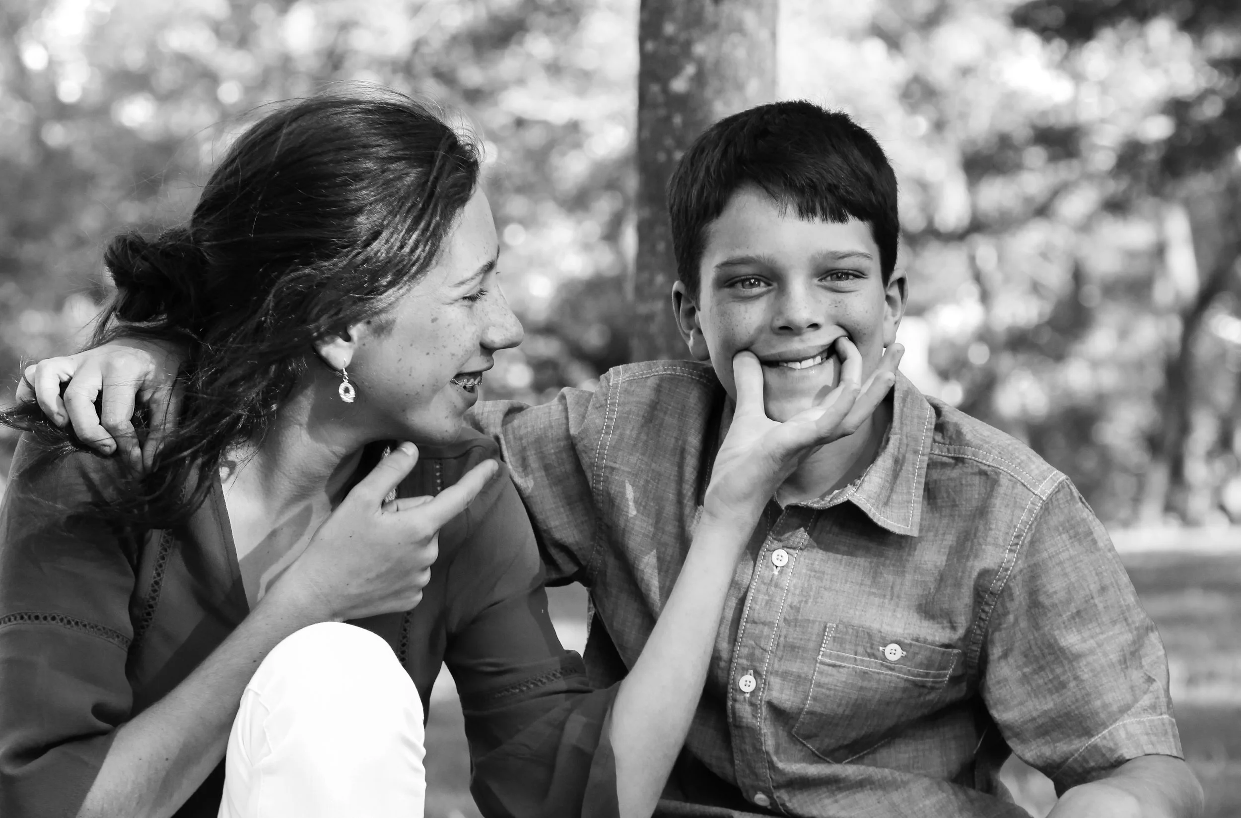 A woman and a boy are outdoors, smiling and having fun together. The woman is sitting close and whispering into the boy's ear while he playfully holds his fingers against his face. The background shows blurred trees.