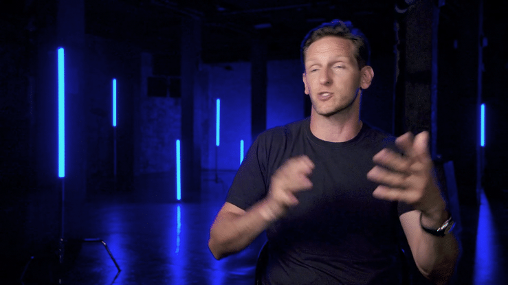 A man with short hair and wearing a black t-shirt is sitting in a dark room with blue neon lights, talking and gesturing with his hands.