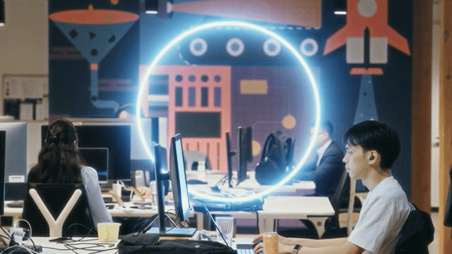 Office workers at desks with computers in front of a wall mural featuring science and technology-themed graphics and neon lighting.