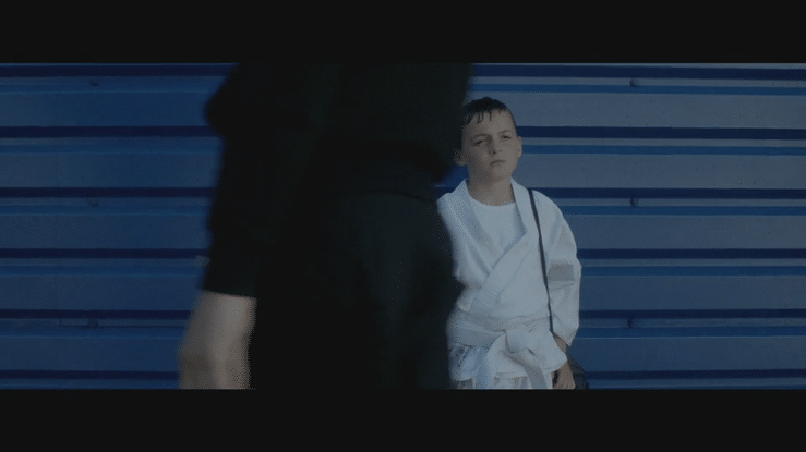 A young boy in a white judo uniform sitting against a blue wall, looking up at an adult standing in front of him.