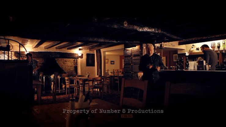 Interior of a cozy restaurant or pub with wooden furniture, a bar area, and a fireplace, dimly lit with warm lighting, featuring a man standing at the bar and other patrons seated at tables.