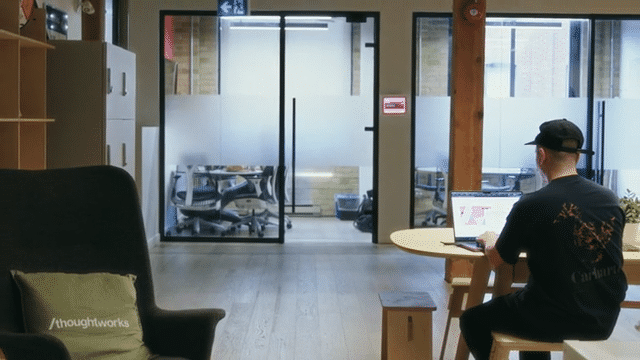 Person sitting at a round desk working on a laptop in a modern office with glass doors and chairs in the background.