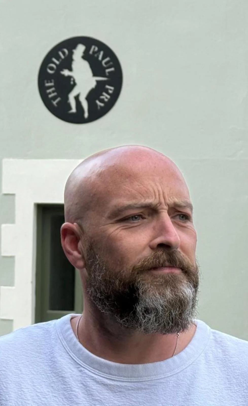 A bald man with a beard and a serious expression standing outside near a white building with a black circular logo that reads 'The Old Paul Peer'.