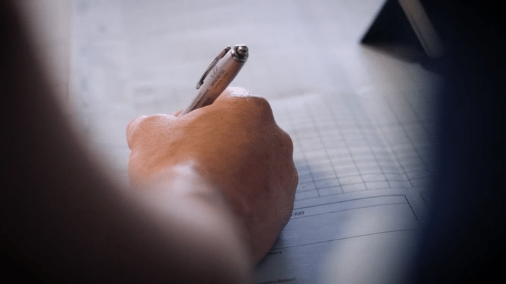 A person's hand holding a pen and writing on a lined notebook with a grid section, on a desk.