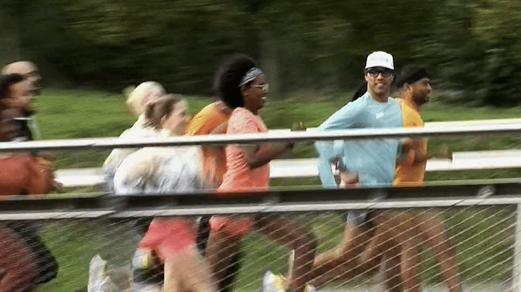 Group of people running outdoors, some wearing athletic clothes and sunglasses, running along a metal railing with greenery in the background.