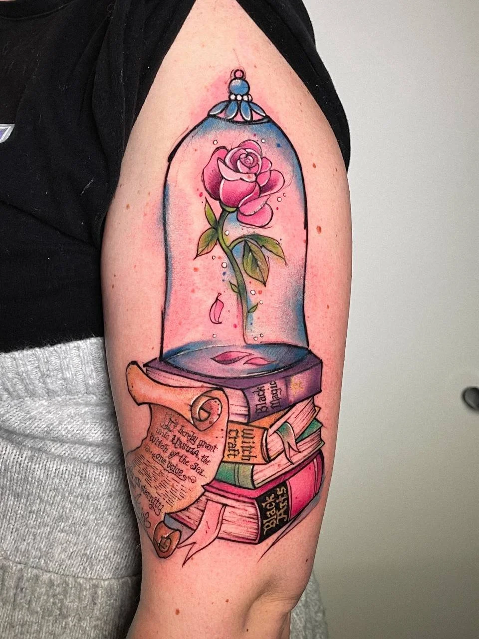 Disney-inspired color tattoo with a rose under a glass dome and books on the upper arm by Maya Marina Tattoo.