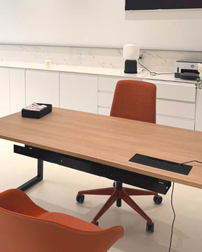 Salle de réunion avec une table en bois, deux chaises orange et un mur blanc avec comptoir, lampe et imprimante.