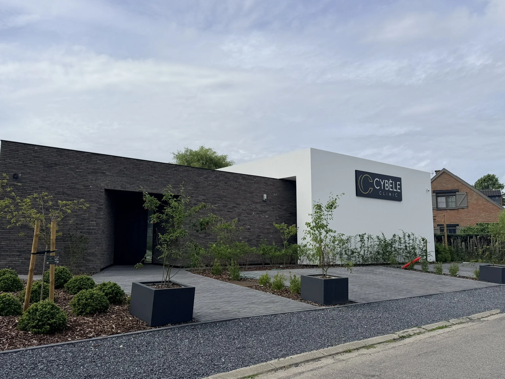 Vue de l'extérieur d'une clinique appelée Cybele Clinic avec un bâtiment en brique foncée, un mur blanc, un parking pavé et des arbres et arbustes dans des jardinières.