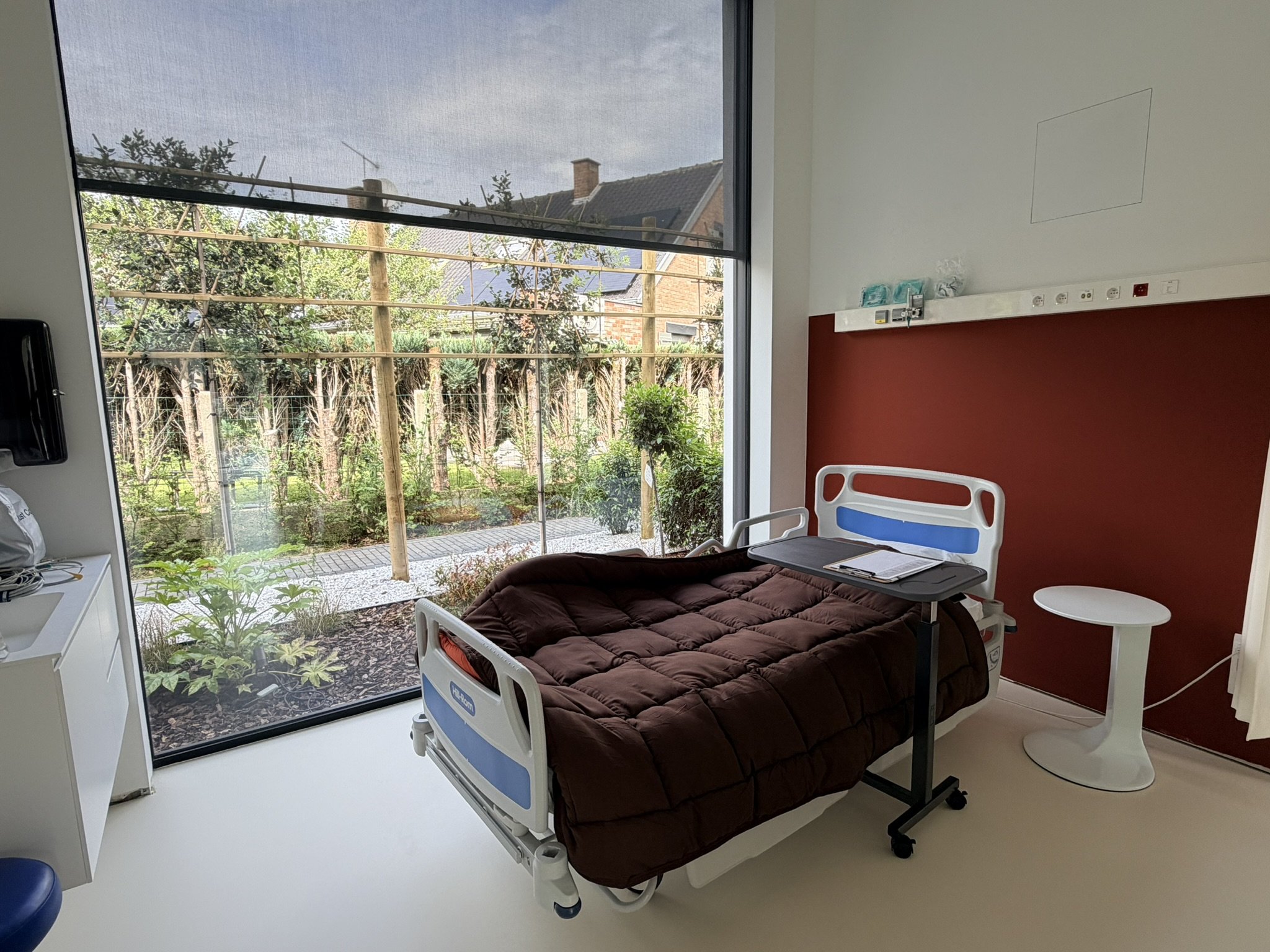 Une chambre d'hôpital avec un lit d'hôpital équipé en matelas brun, une table d'appoint blanche, et une fenêtre large laissant entrer la lumière naturelle, avec une vue sur un jardin extérieur zen