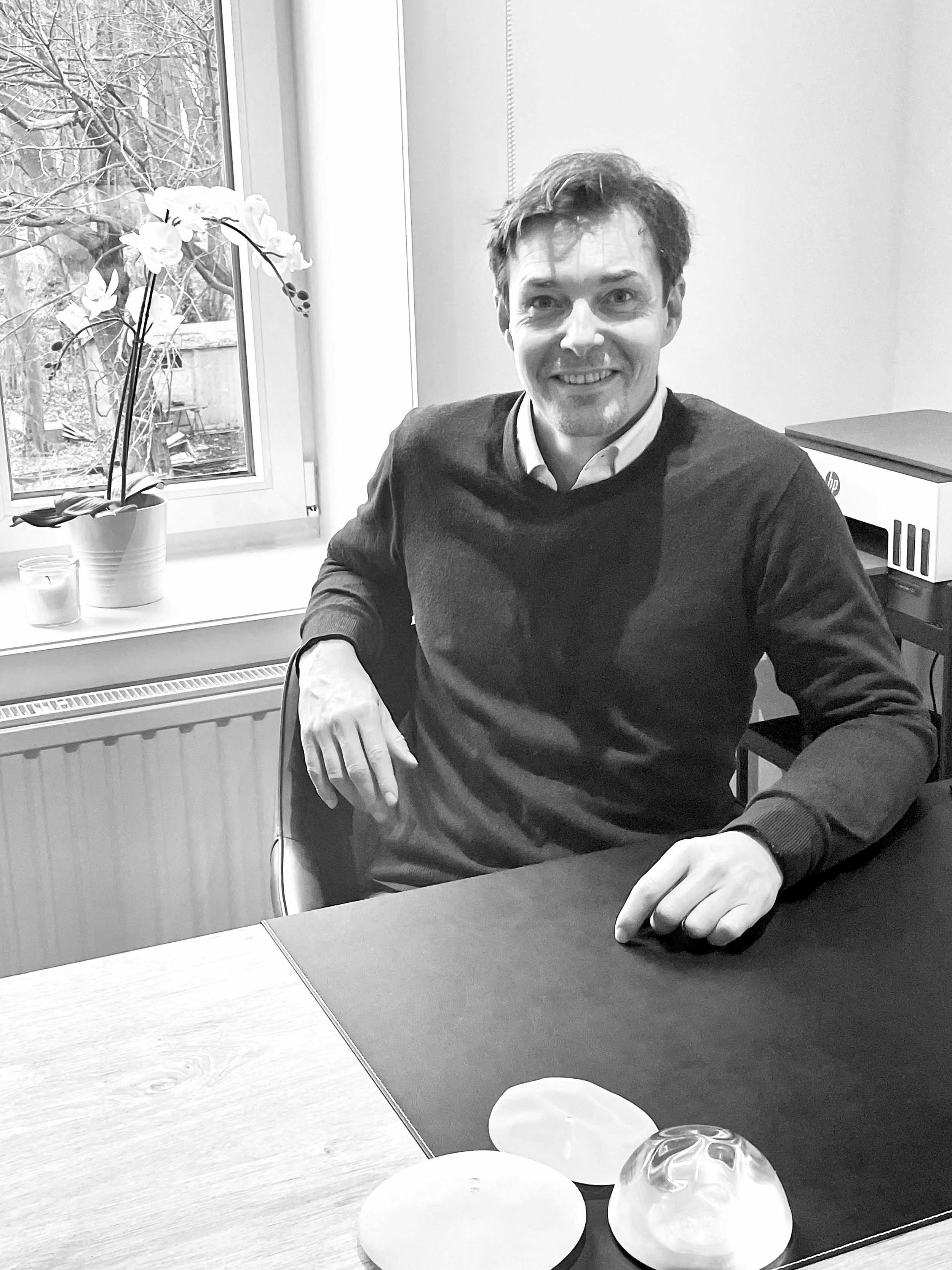 Le Dr Raphaël Gheerardyn, assis à une table en intérieur, avec une fenêtre derrière lui montrant du gel et des branches d'arbres. Sur la table, il y a des objets décoratifs en forme de bulbes ou de coquillages. La scène est en noir et blanc.