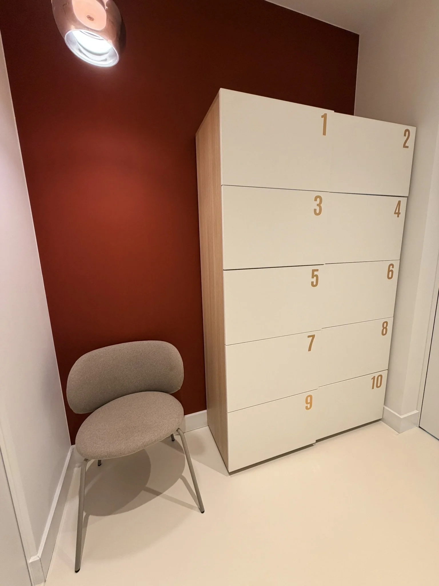 Une chaise beige avec un dossier courbé et quatre jambes métalliques se trouve à côté d'une grande armoire blanche numérotée de 1 à 10 sur la porte. La pièce possède un mur rouge et un plafond avec un plafonnier cylindrique enroulé de tissu.