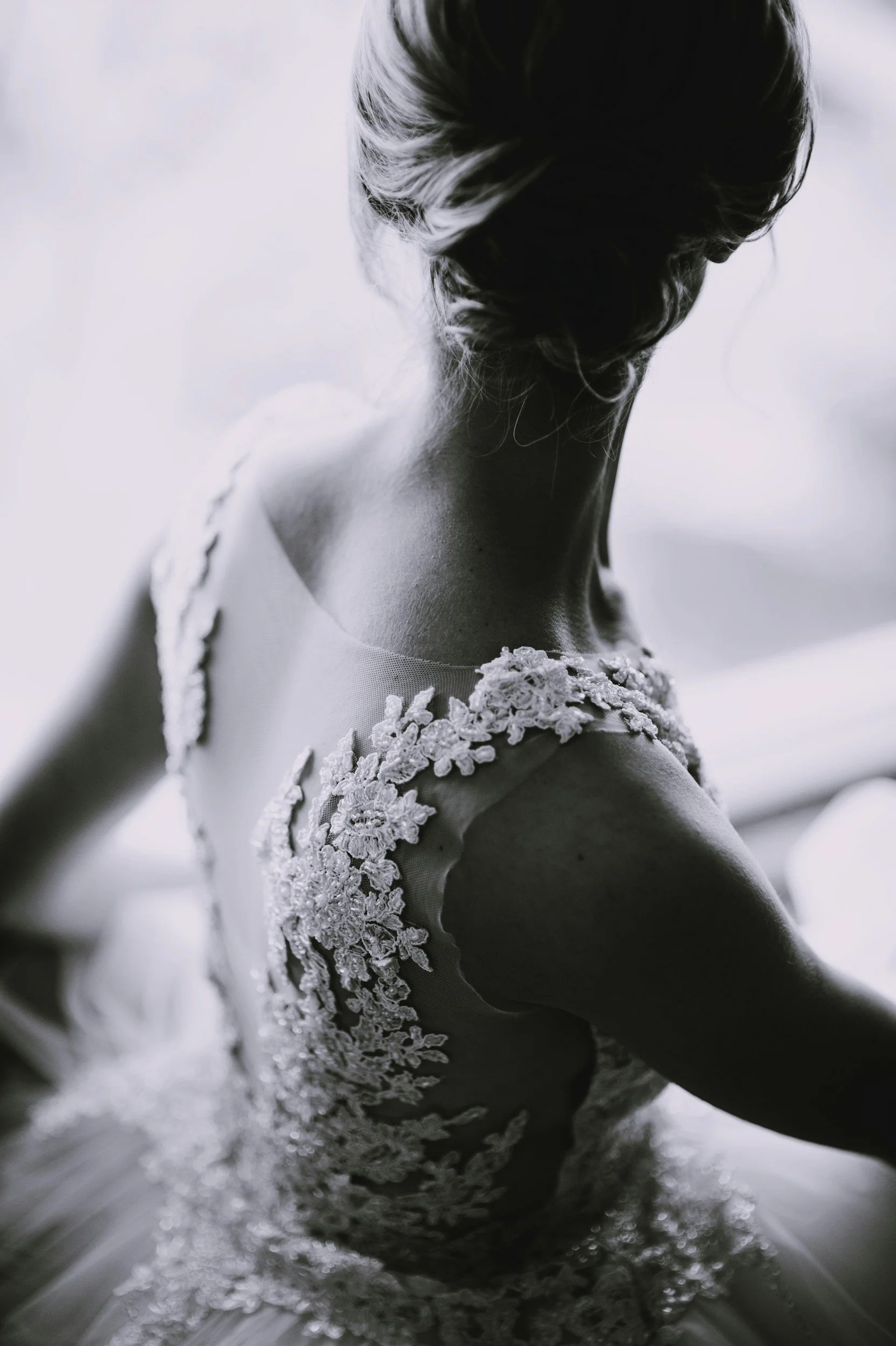 A woman in a wedding dress with floral lace details, photographed from behind in black and white.