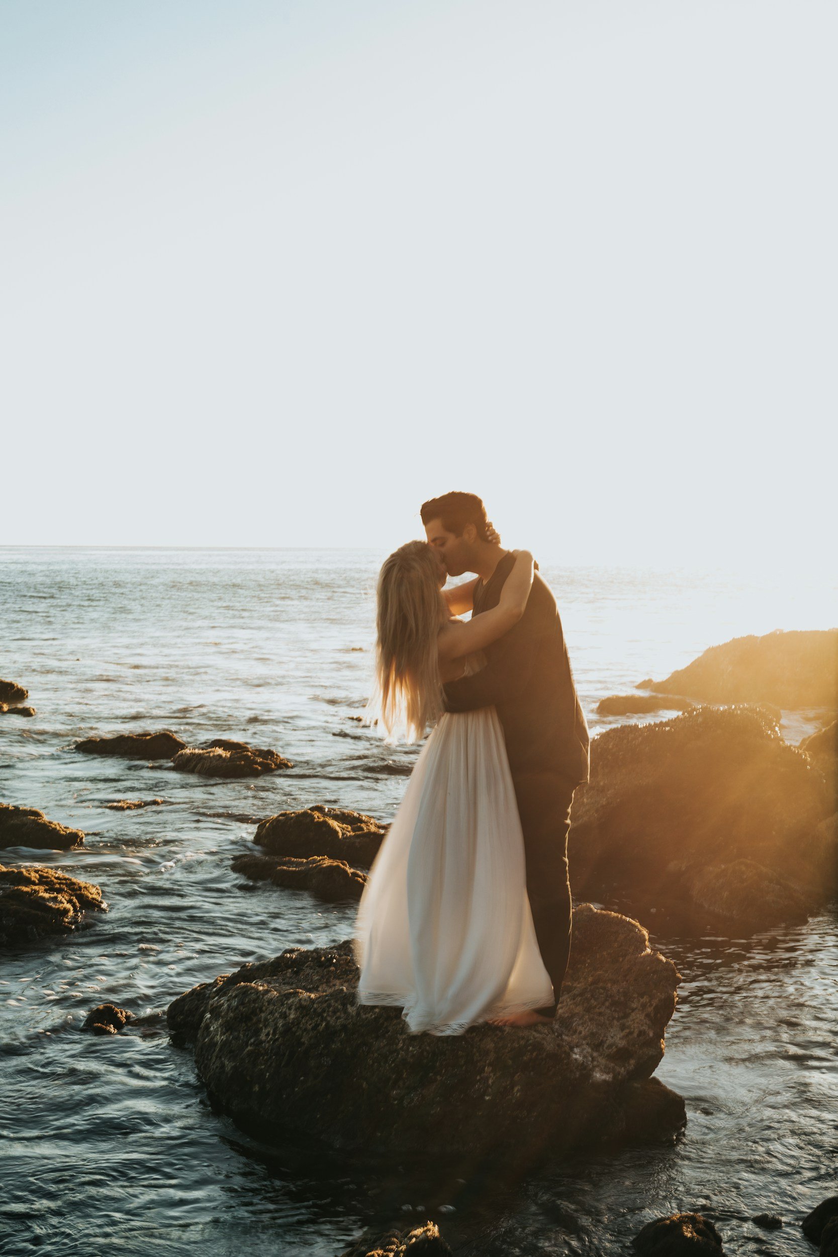 A couple stands on rocks by the ocean at sunset, sharing a kiss, with the woman wearing a white dress and the man in dark clothing.