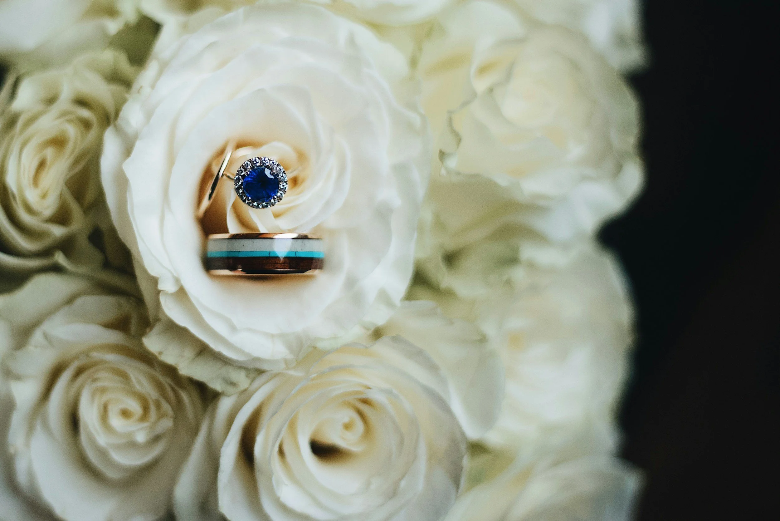 A close-up of a diamond ring with a large blue gemstone surrounded by smaller diamonds, resting on a white rose surrounded by cream-colored roses.