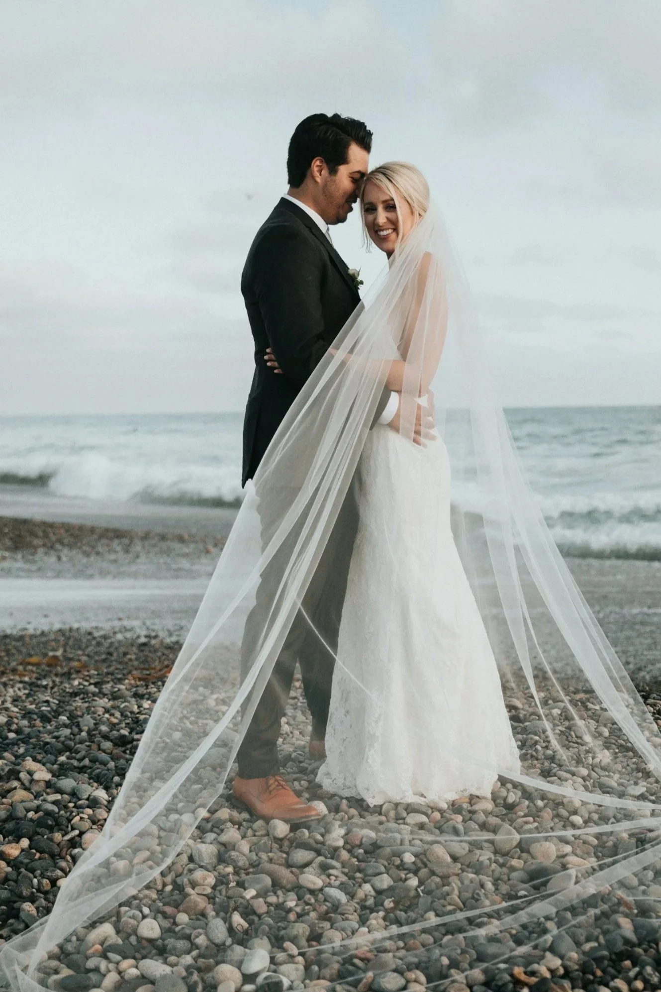A couple dressed in wedding attire standing on a rocky beach with the ocean in the background, sharing an intimate moment.