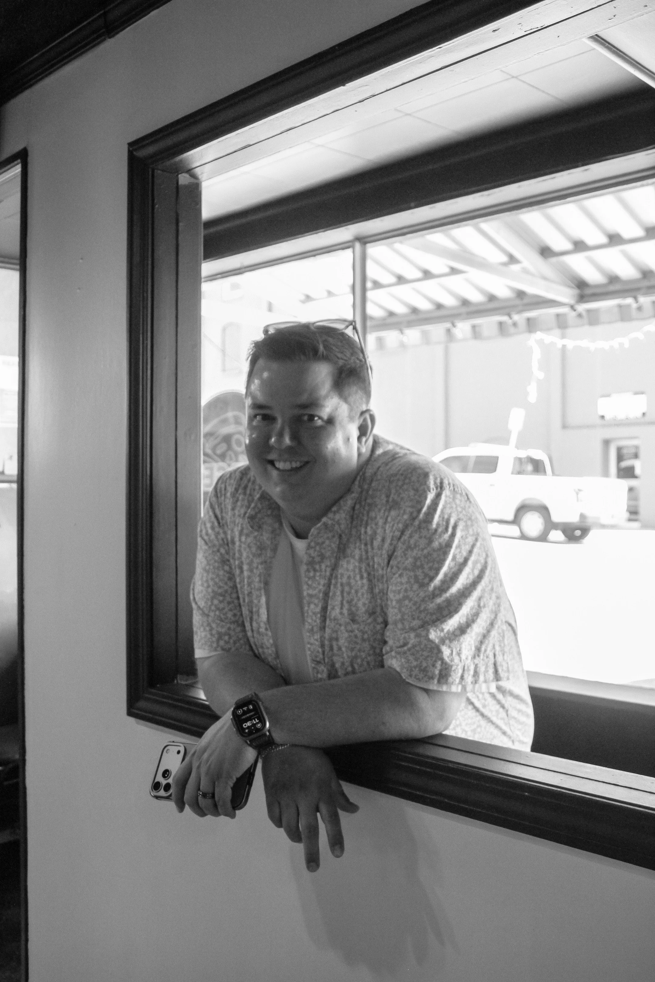 A smiling young man with glasses on his head is leaning out of a window, holding a smartphone in his right hand which is resting on his knee. He is wearing a patterned shirt and a smartwatch on his left wrist. The scene is in black and white, with an exterior car visible in the background.