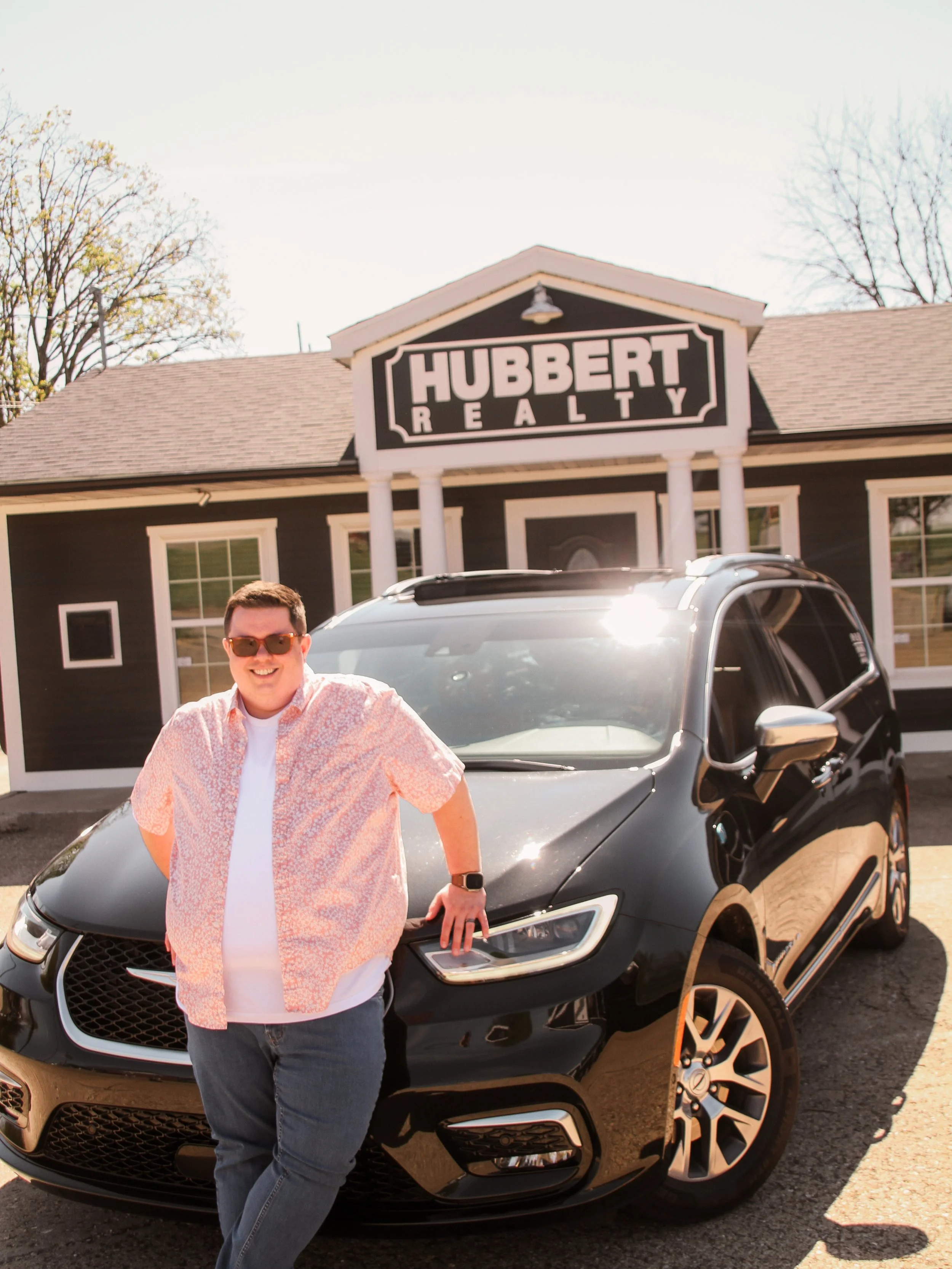 Young man with sunglasses smiling, standing next to a black SUV in front of a real estate office named Hubbert Realty, in a sunny outdoor setting.