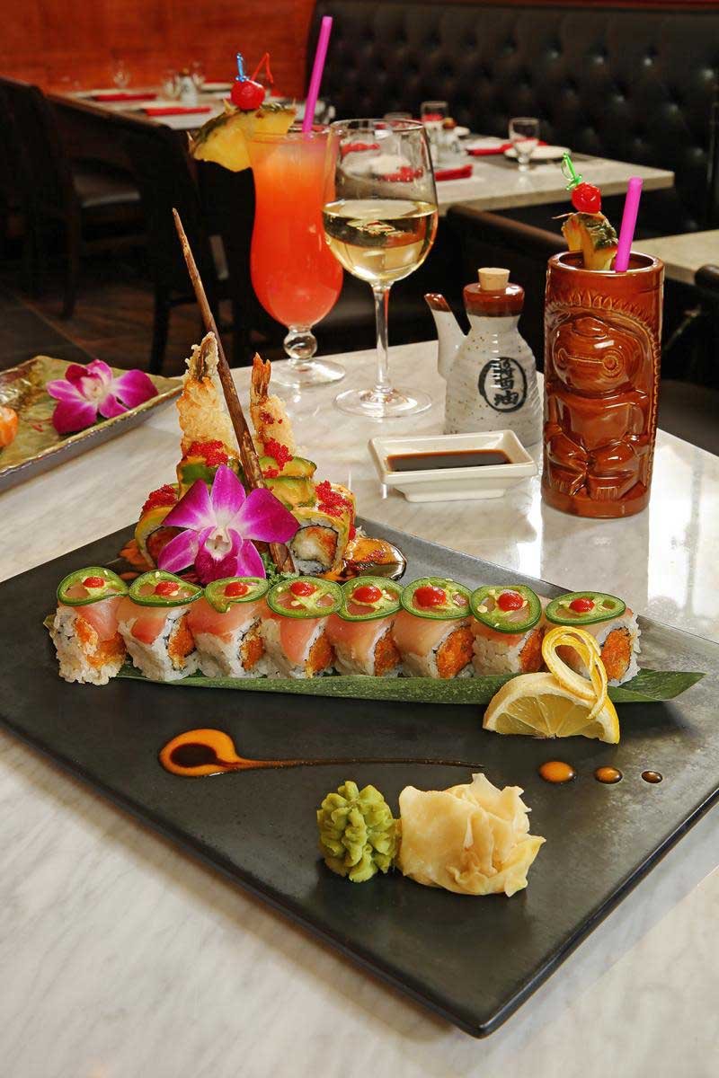 Plate of sushi rolls garnished with slices of jalapeño and a lime wedge, with wasabi and ginger on the side, served on a black rectangular plate in a restaurant setting.