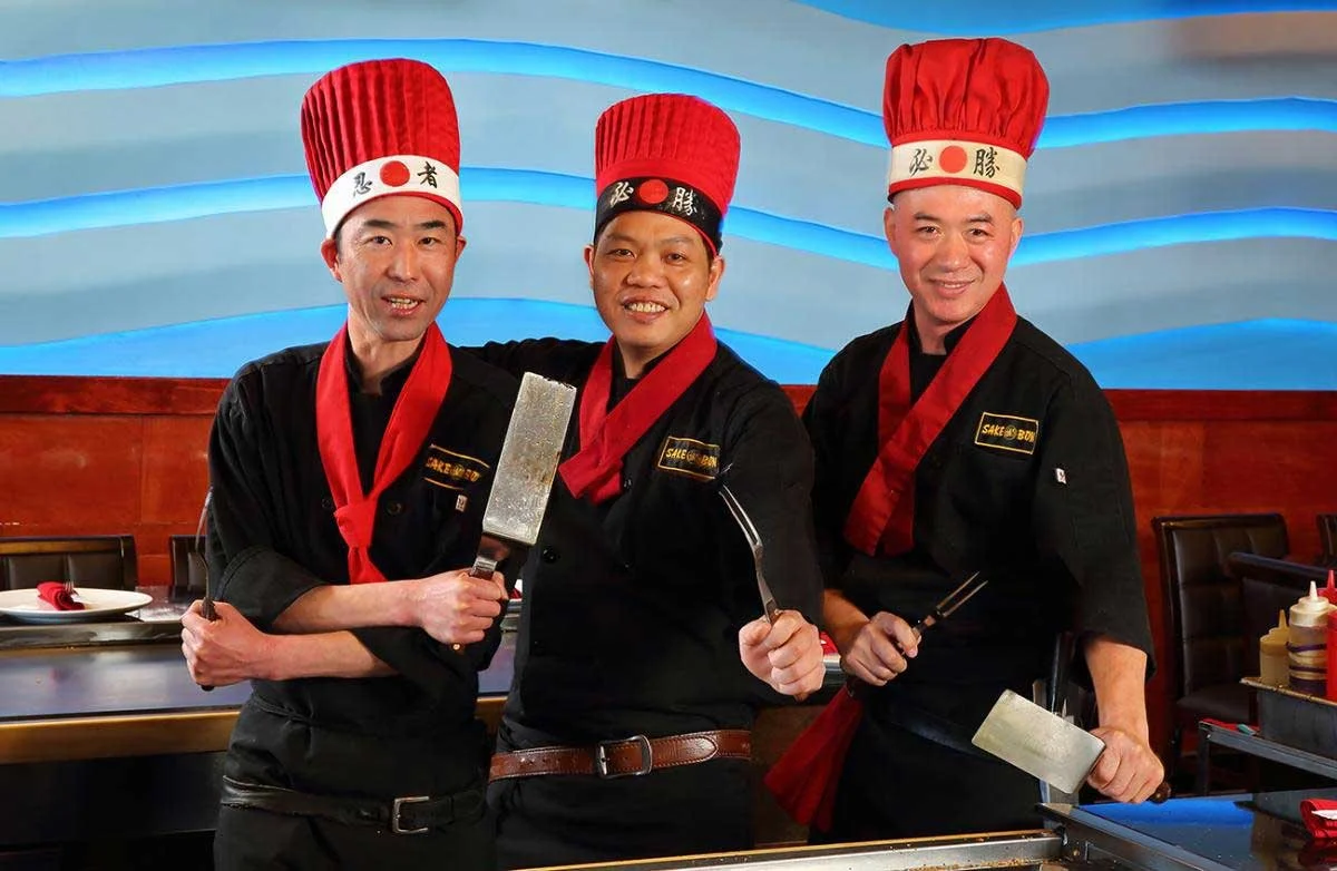 Three male sushi chefs in black uniforms and red hats standing in a restaurant kitchen, smiling and holding cooking utensils.
