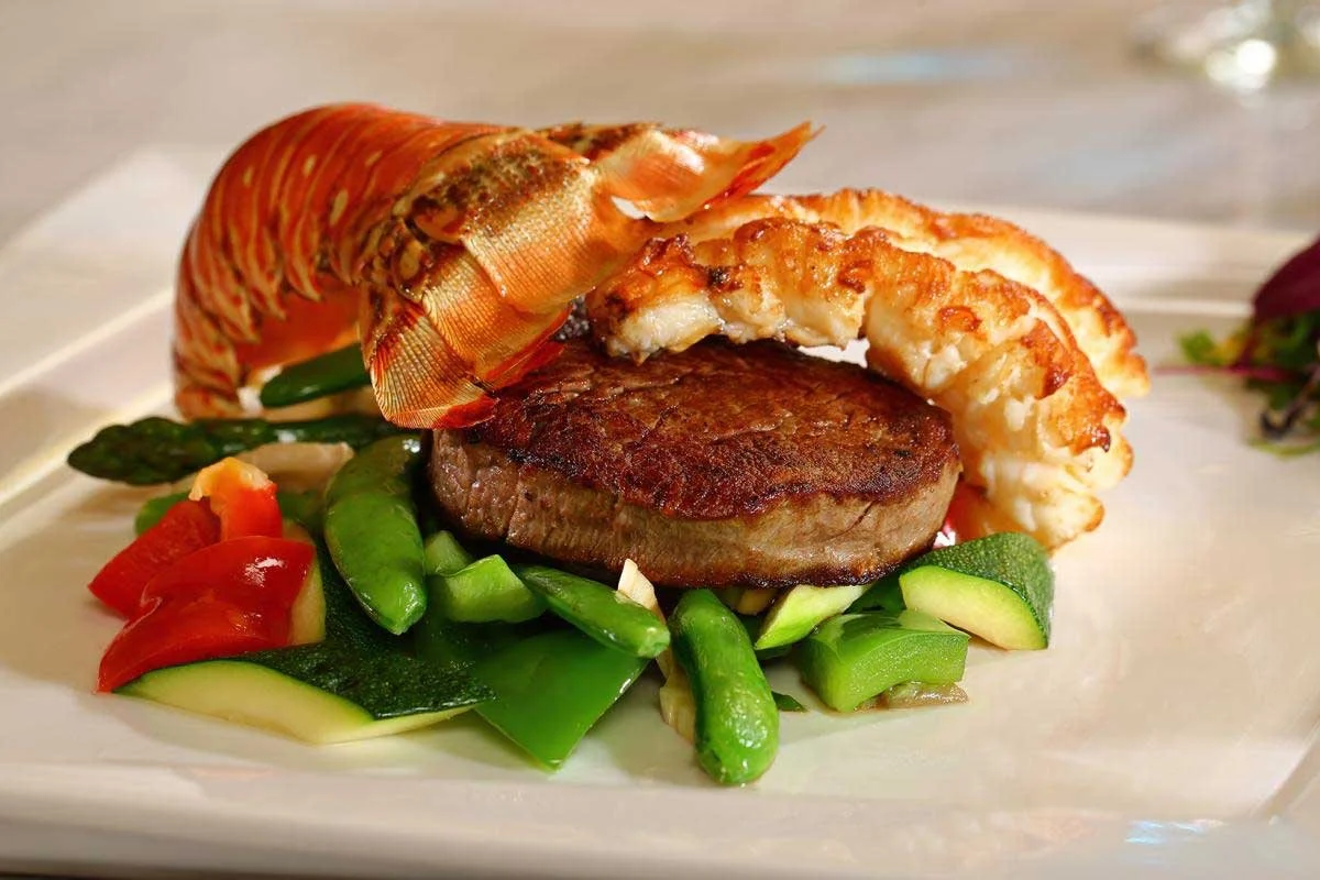 A plate with a lobster tail, a cooked steak, and assorted vegetables including asparagus, zucchini, and tomatoes.