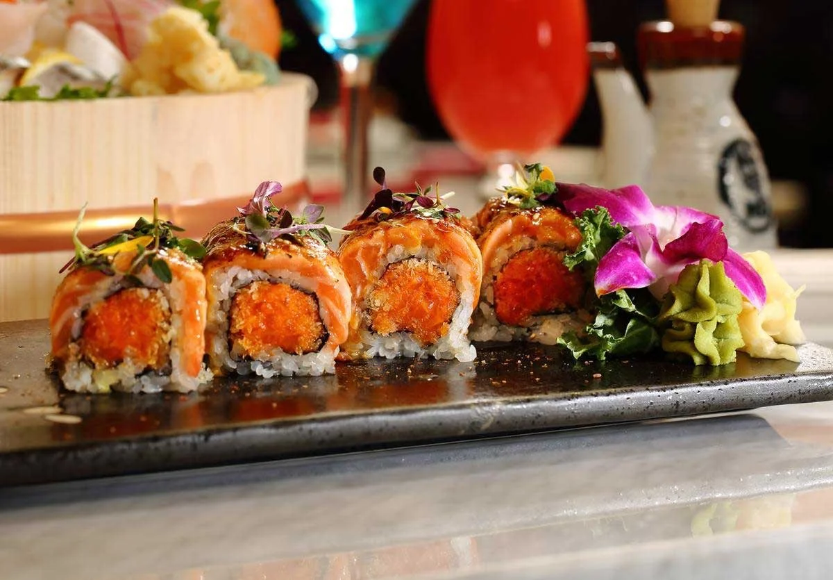 Close-up of a plate with four pieces of sushi rolls garnished with microgreens, served with wasabi, pickled ginger, and edible purple flowers on the side, with a blurred background of drinks and condiments.
