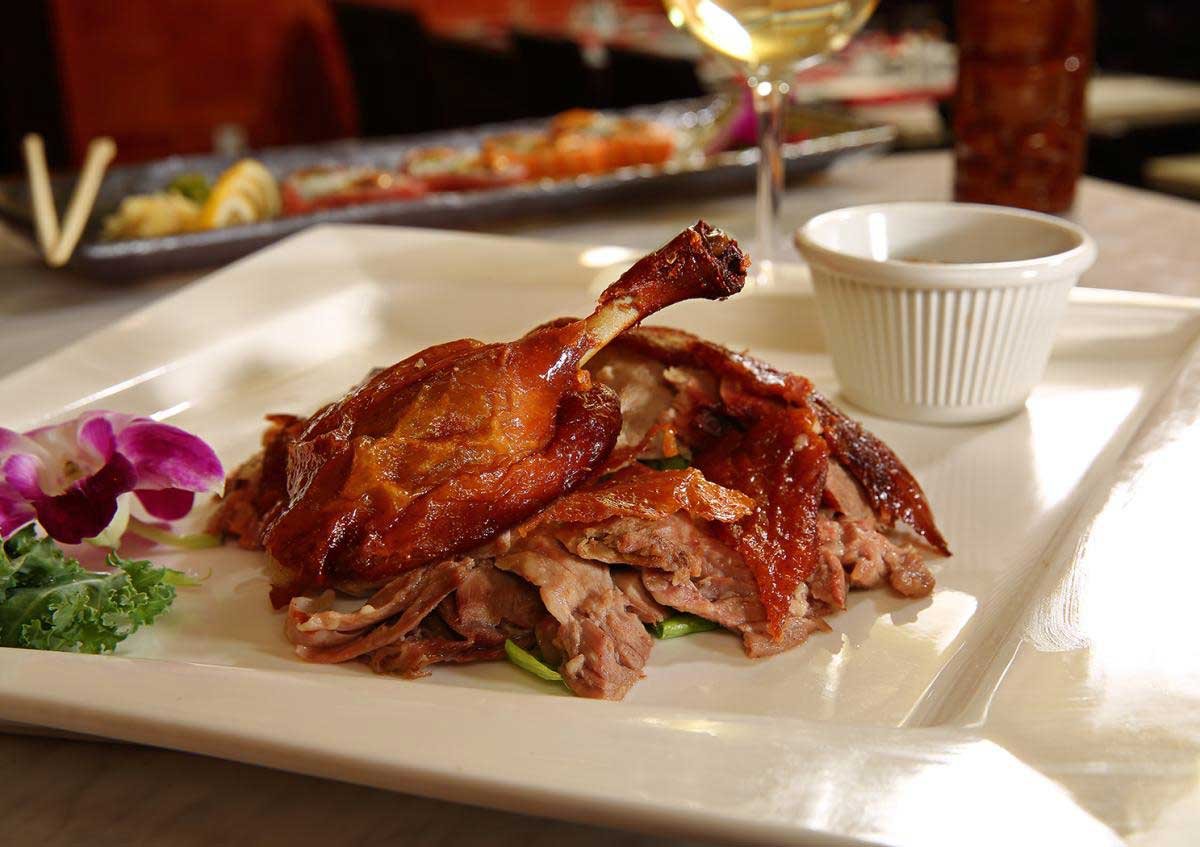 A plate with roast meat and a crispy chicken leg, garnished with purple flowers and greens, with a small ramekin and a wine glass in the background.
