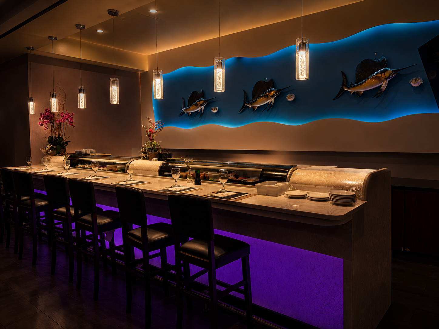 Interior of a modern sushi restaurant with a sushi bar, black chairs, and decorative ocean-themed wall art featuring marlins and a blue wave backdrop, illuminated with pendant and purple accent lighting.