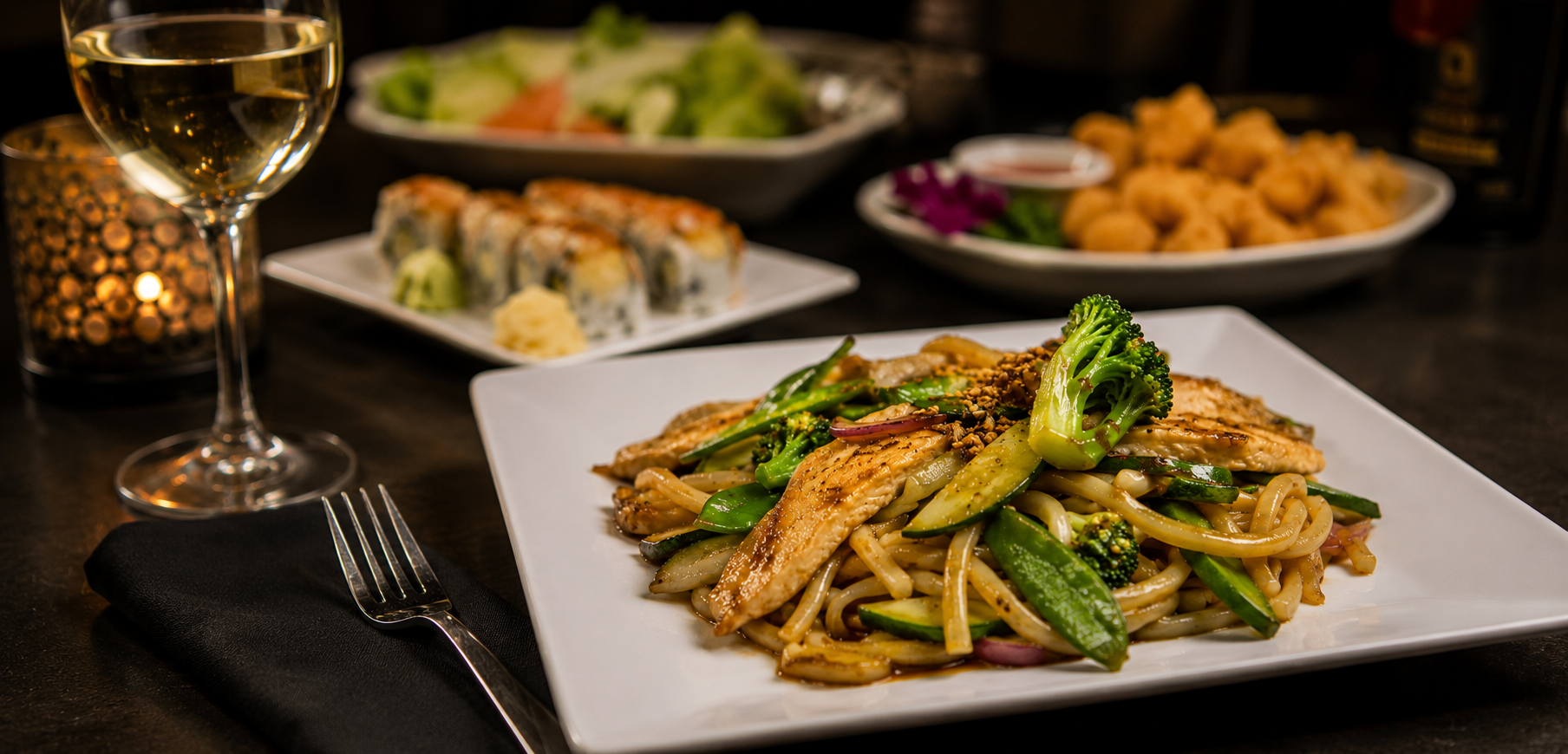 A plate of stir-fried noodles with vegetables and chicken, accompanied by sushi rolls, fried shrimp, a glass of white wine, and a lit candle on a dark table.