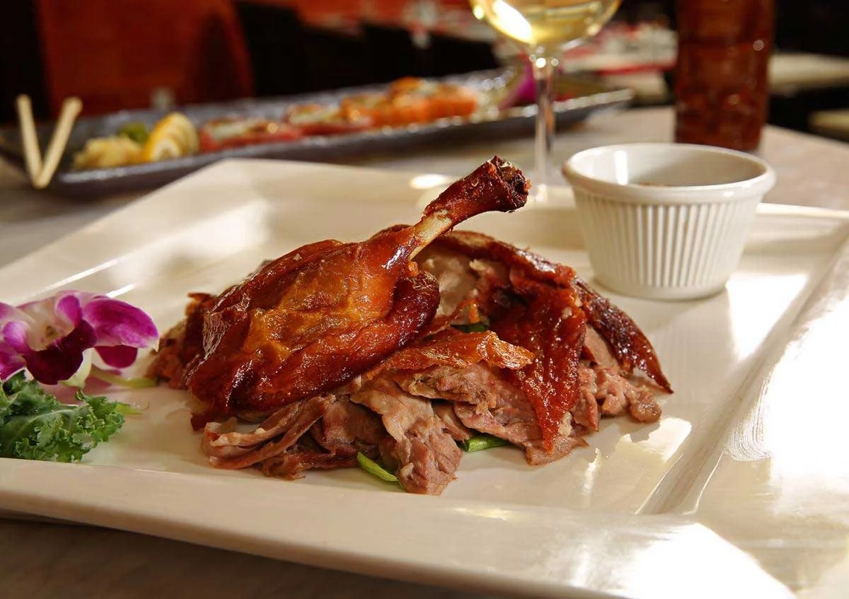 A plate with roasted duck, sliced meat, purple flowers, greens, a small white ramekin, and a glass of white wine.