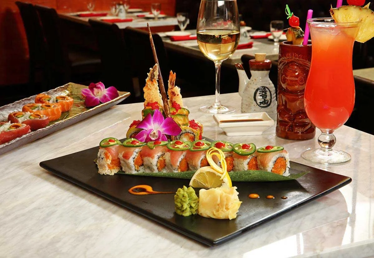 A plate of sushi rolls garnished with slices of jalapeno, a lemon wedge, wasabi, and ginger on a black rectangular plate. In the background, there is a glass of white wine and a colorful cocktail with a pineapple slice, pineapple chunks, and a cherry