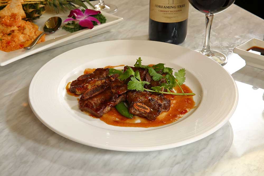 Plate of braised short ribs garnished with cilantro and served with sauce, with a glass of red wine and a bottle of wine in the background.