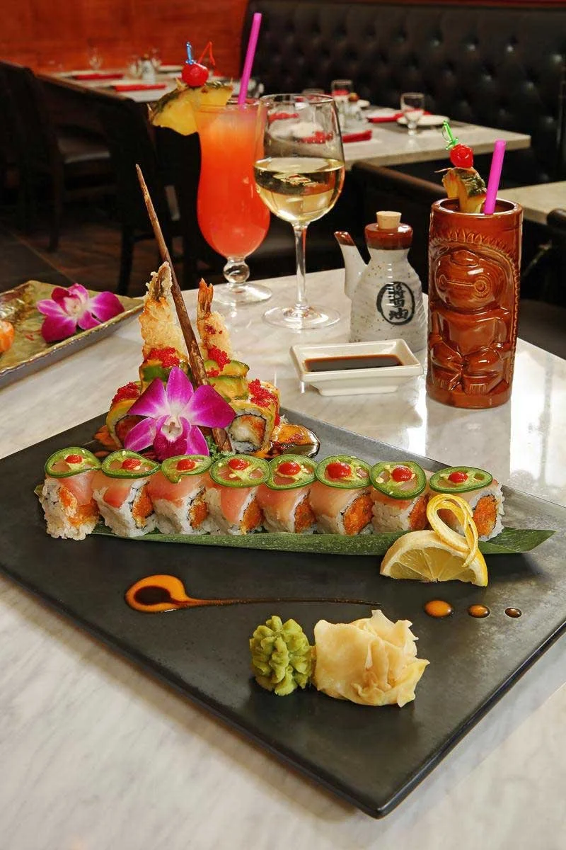 A plate of assorted sushi rolls garnished with green chili slices and red sauce, accompanied by a lemon wedge, wasabi, and pickled ginger, served on a black rectangular plate with decorative sauce, on a white marble table. In the background, there ar