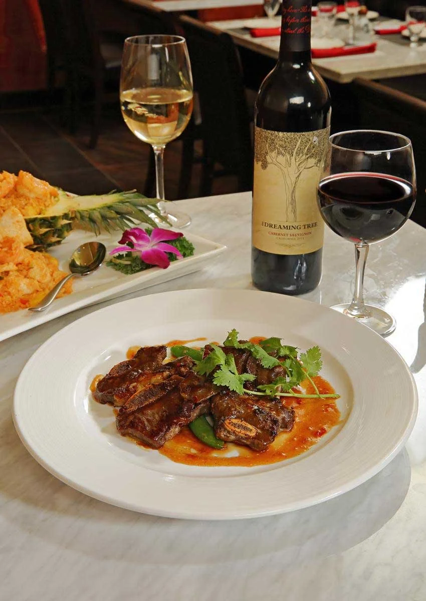 A white plate with braised beef ribs garnished with cilantro on a tomato-based sauce. Two glasses of red and white wine and a bottle of Cabernet Sauvignon are on the table. In the background, there is a platter of fruit and a pineapple with a flower 