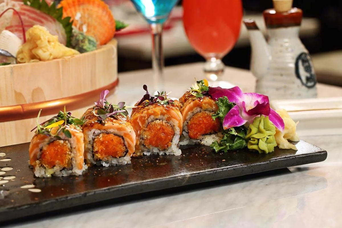 Sushi roll slices with orange fish, topped with microgreens, served with wasabi, pickled ginger, and garnished with a purple orchid on a black plate.