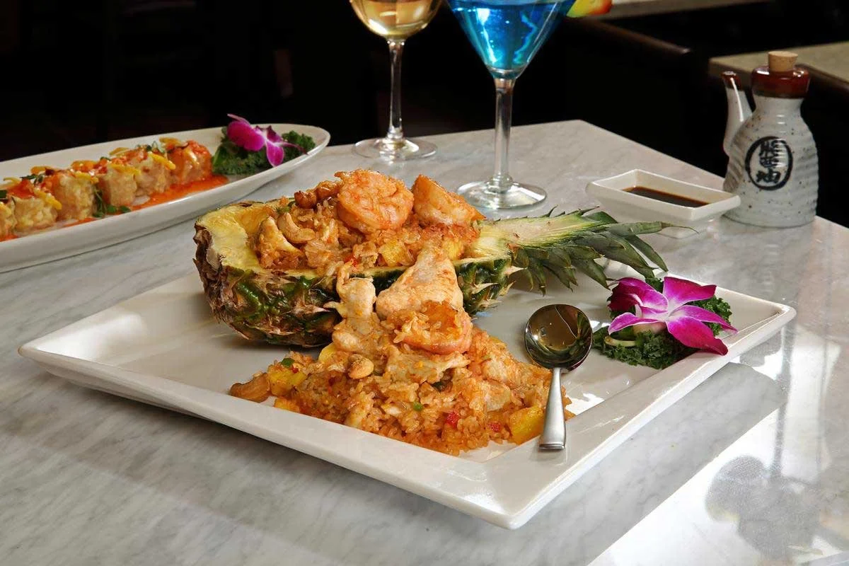 Open pineapple filled with fried rice and shrimp, garnished with tropical flowers, on a white rectangular plate.