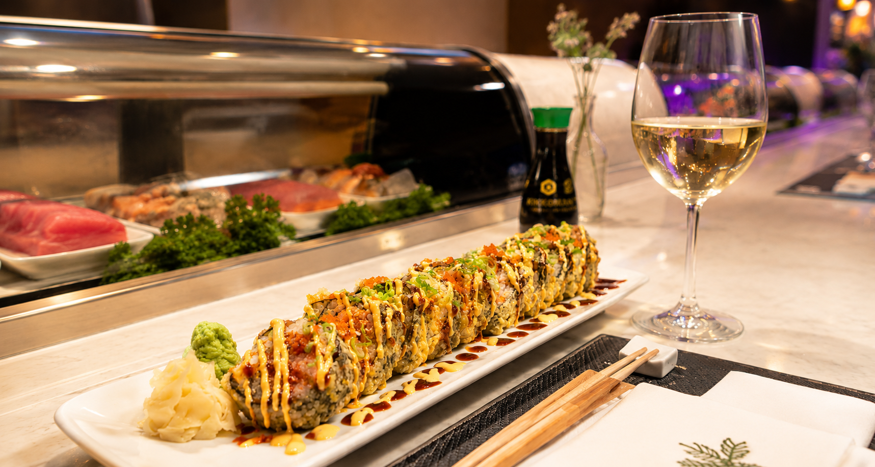 A plate of sushi rolls decorated with sauce and garnished with green onions, served with wasabi and pickled ginger, and a glass of white wine on a restaurant counter, with a sushi display case and condiments in the background.